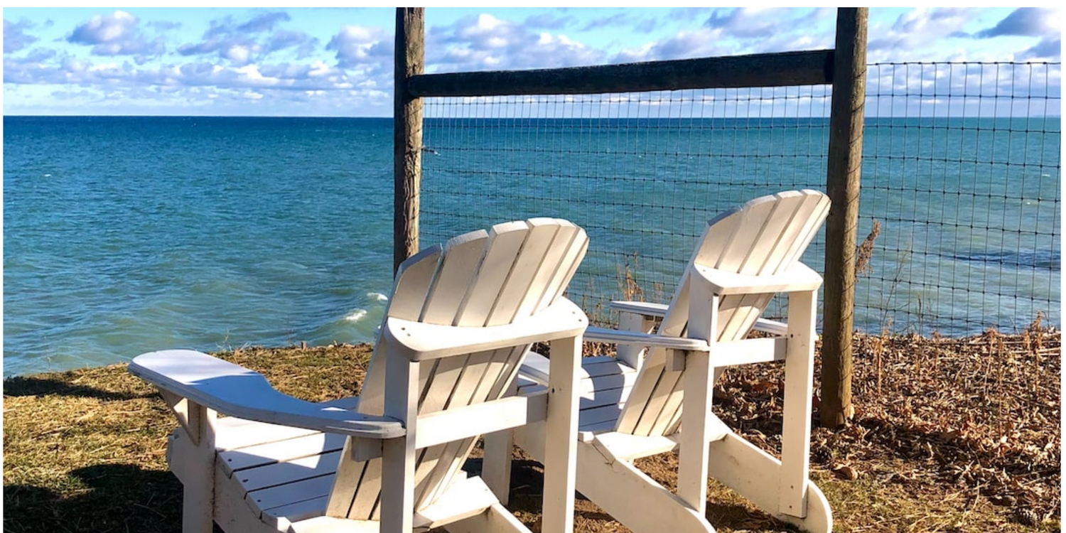 Two white Adirondack chairs overlooking a beautiful blue ocean, set against a rustic wooden fence—perfect for a relaxing outdoor retreat.